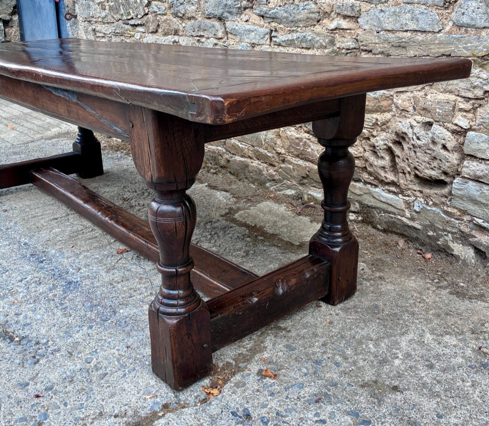 A long dark oak refectory dining table featuring a solid plank top, cleated ends, turned legs, and a low stretcher rail. View VI