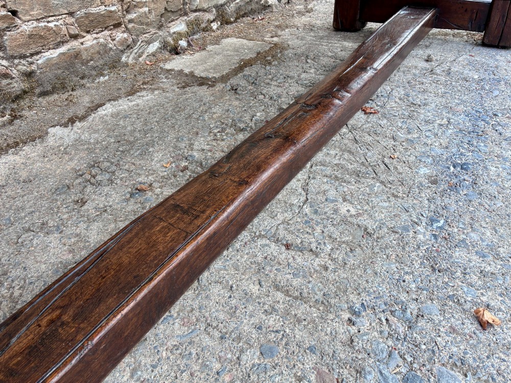 A long dark oak refectory dining table featuring a solid plank top, cleated ends, turned legs, and a low stretcher rail. View VII
