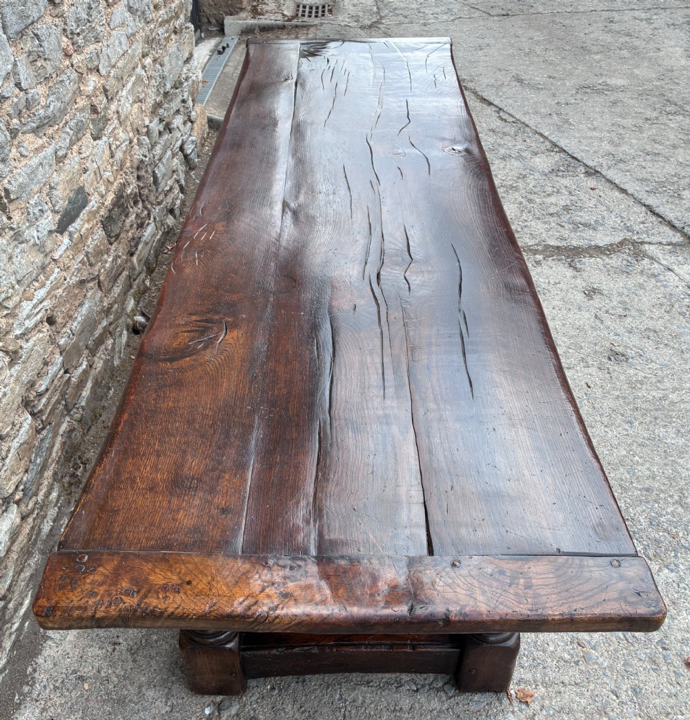 A long dark oak refectory dining table featuring a solid plank top, cleated ends, turned legs, and a low stretcher rail. View IX