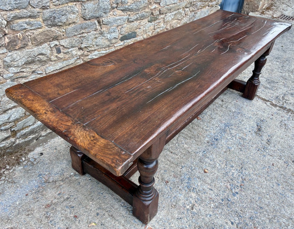 A long dark oak refectory dining table featuring a solid plank top, cleated ends, turned legs, and a low stretcher rail. View X