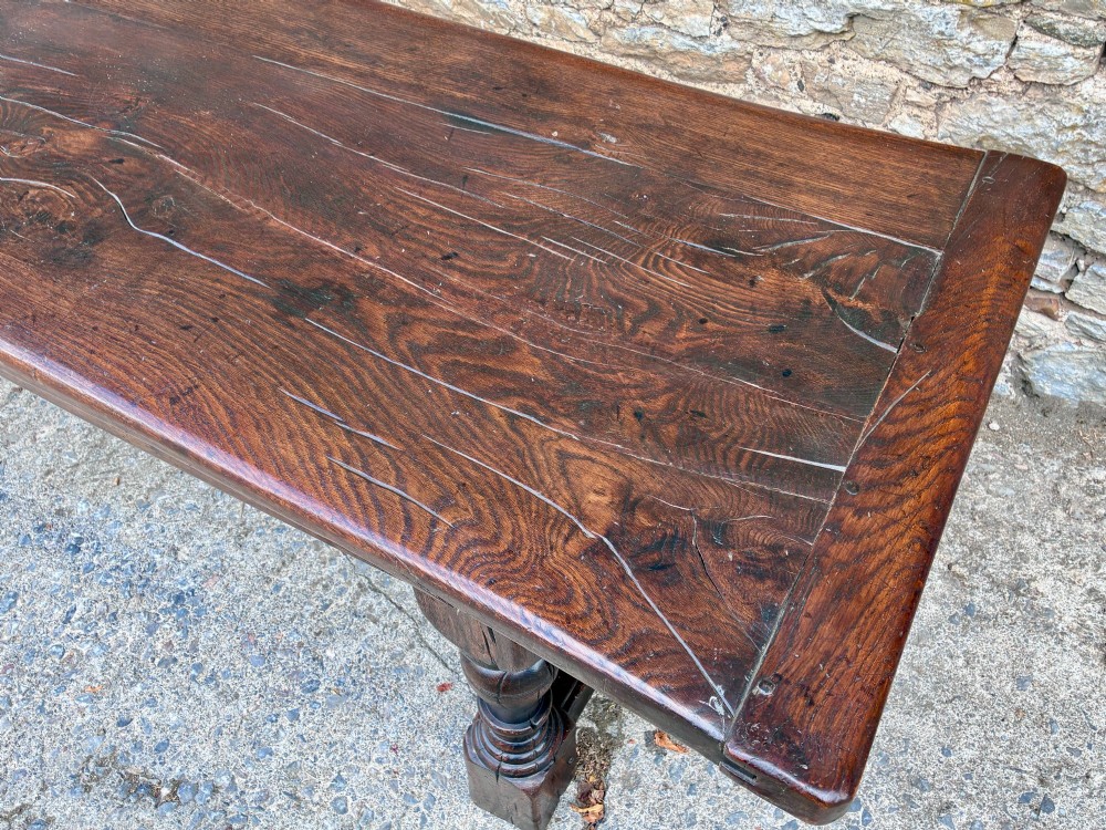 A long dark oak refectory dining table featuring a solid plank top, cleated ends, turned legs, and a low stretcher rail. View XII