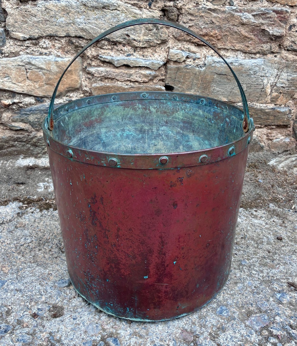 A cylindrical copper bucket featuring a riveted top band, a swing metal handle, and visible green verdigris patination.
