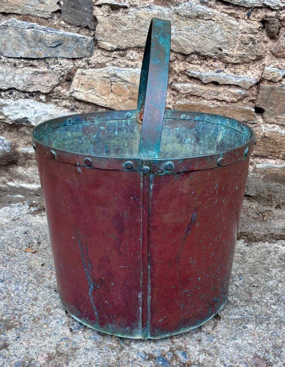 A cylindrical copper bucket featuring a riveted top band, a swing metal handle, and visible green verdigris patination. View V