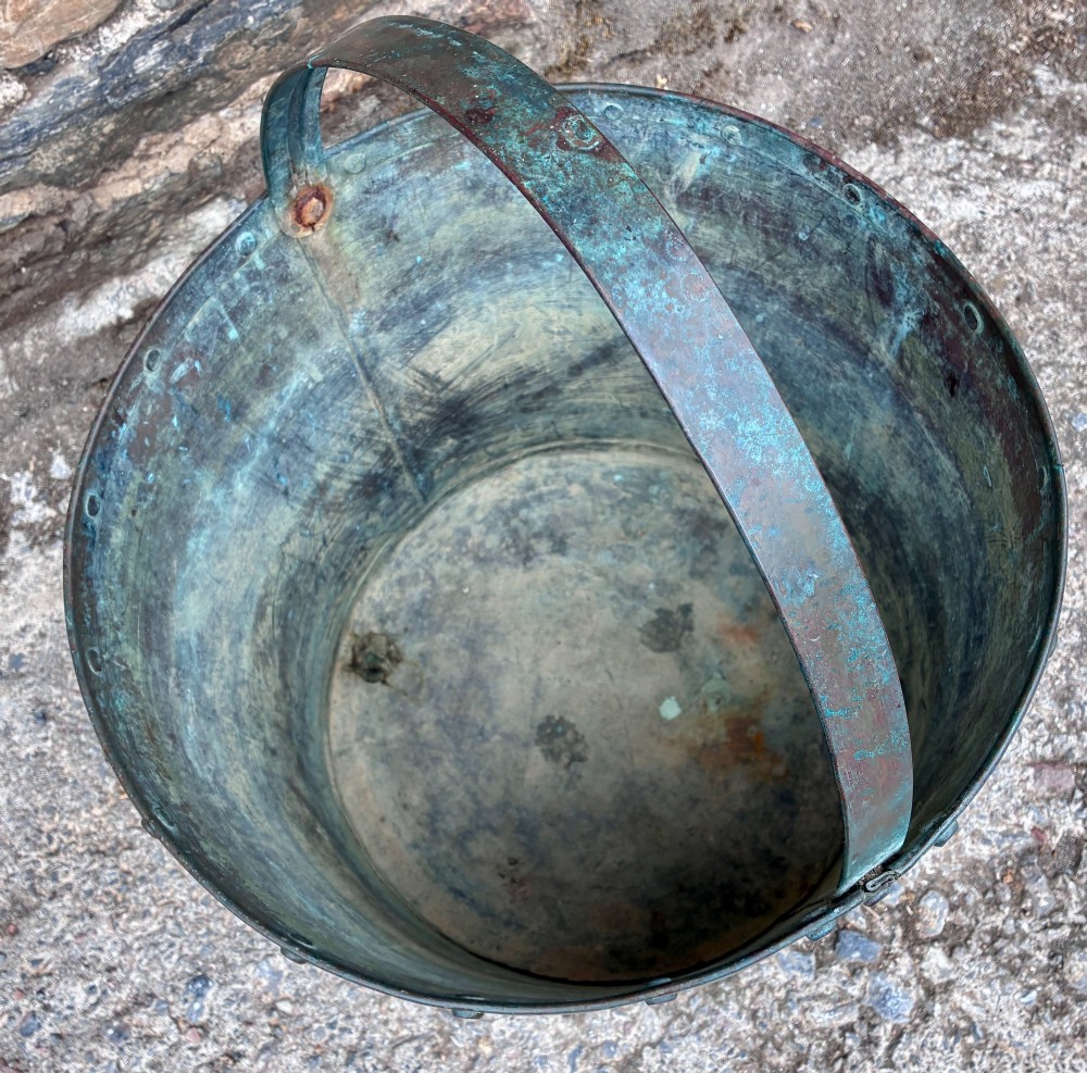 A cylindrical copper bucket featuring a riveted top band, a swing metal handle, and visible green verdigris patination. View VI