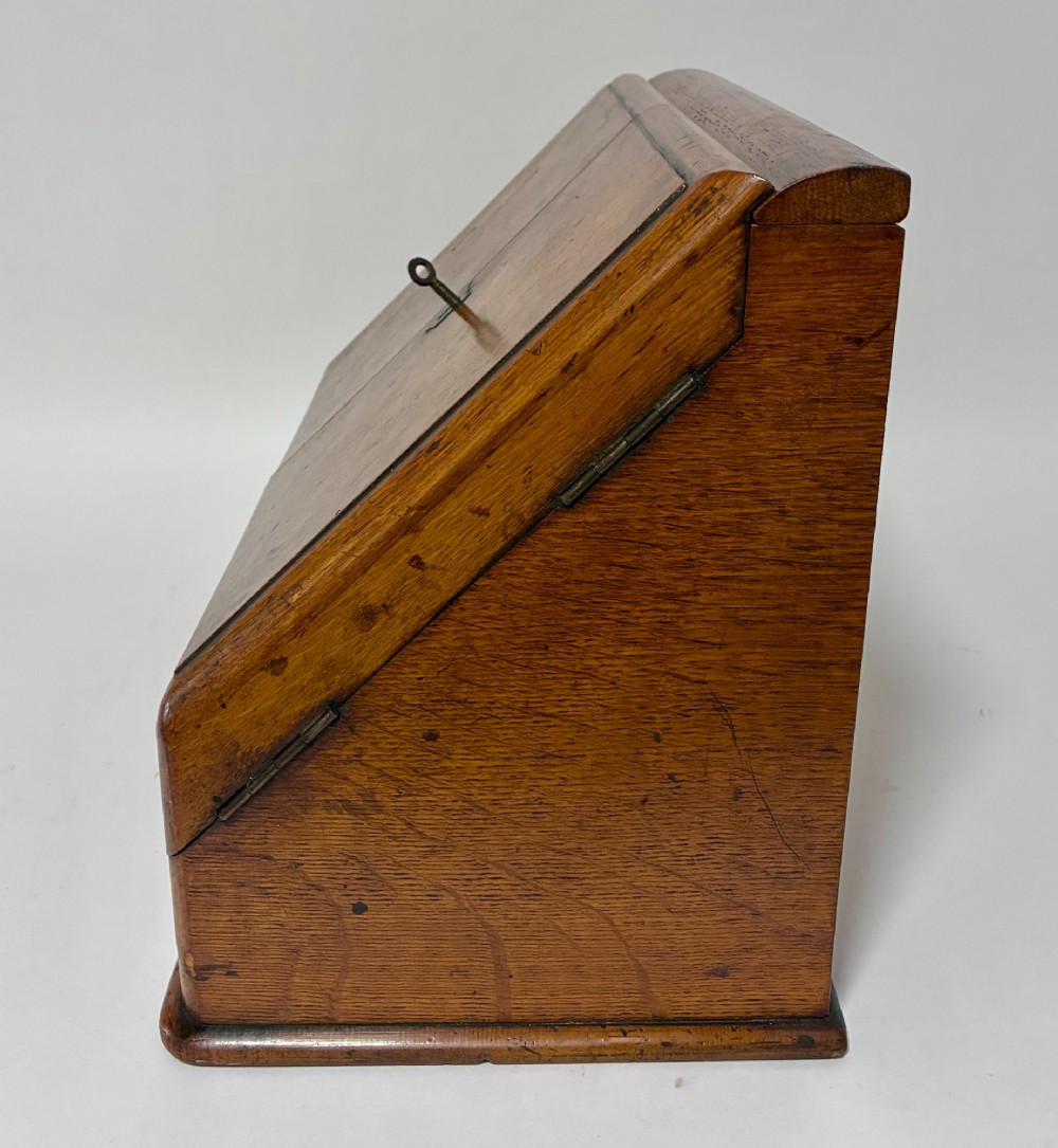 A Victorian oak stationery cabinet featuring sloped double doors, a metal escutcheon with a key, and a moulded plinth base. View IX