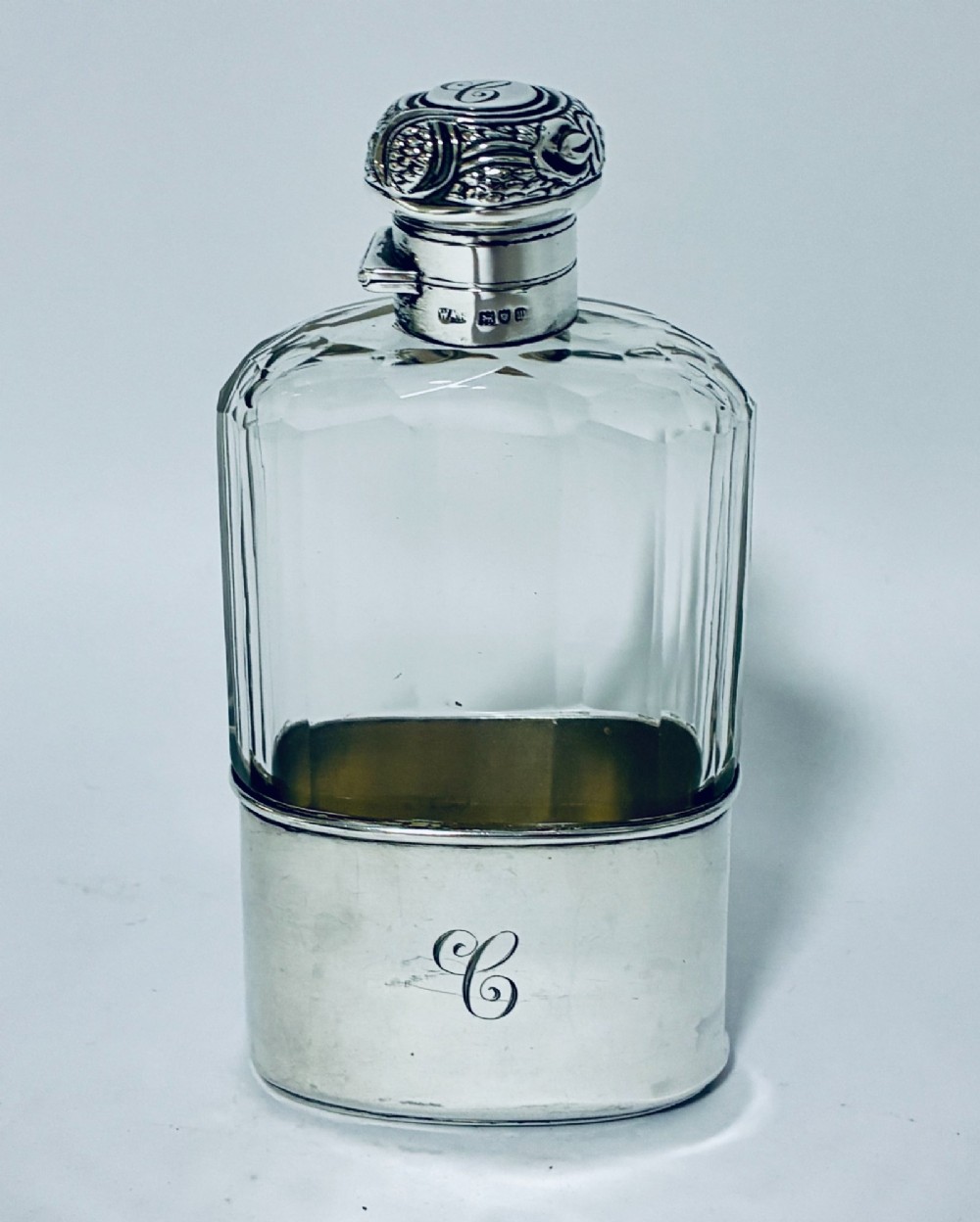 An antique cut-glass hip flask featuring a hallmarked sterling silver collar, chased cap, and a removable silver base cup engraved with the letter C.
