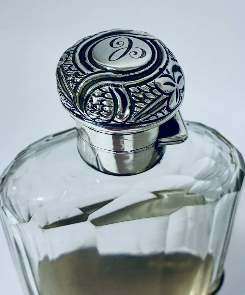 An antique cut-glass hip flask featuring a hallmarked sterling silver collar, chased cap, and a removable silver base cup engraved with the letter C. View IX
