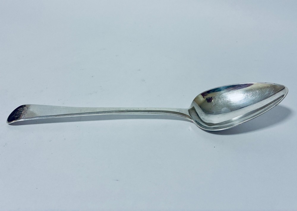 A Georgian sterling silver serving spoon featuring an Old English pattern stem with an engraved crest and a tapered oval bowl against a white backdrop.