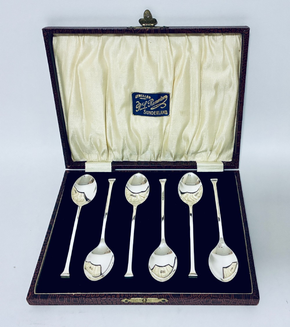 A set of six silver seal top spoons displayed inside a dark presentation box lined with cream satin and navy velvet.