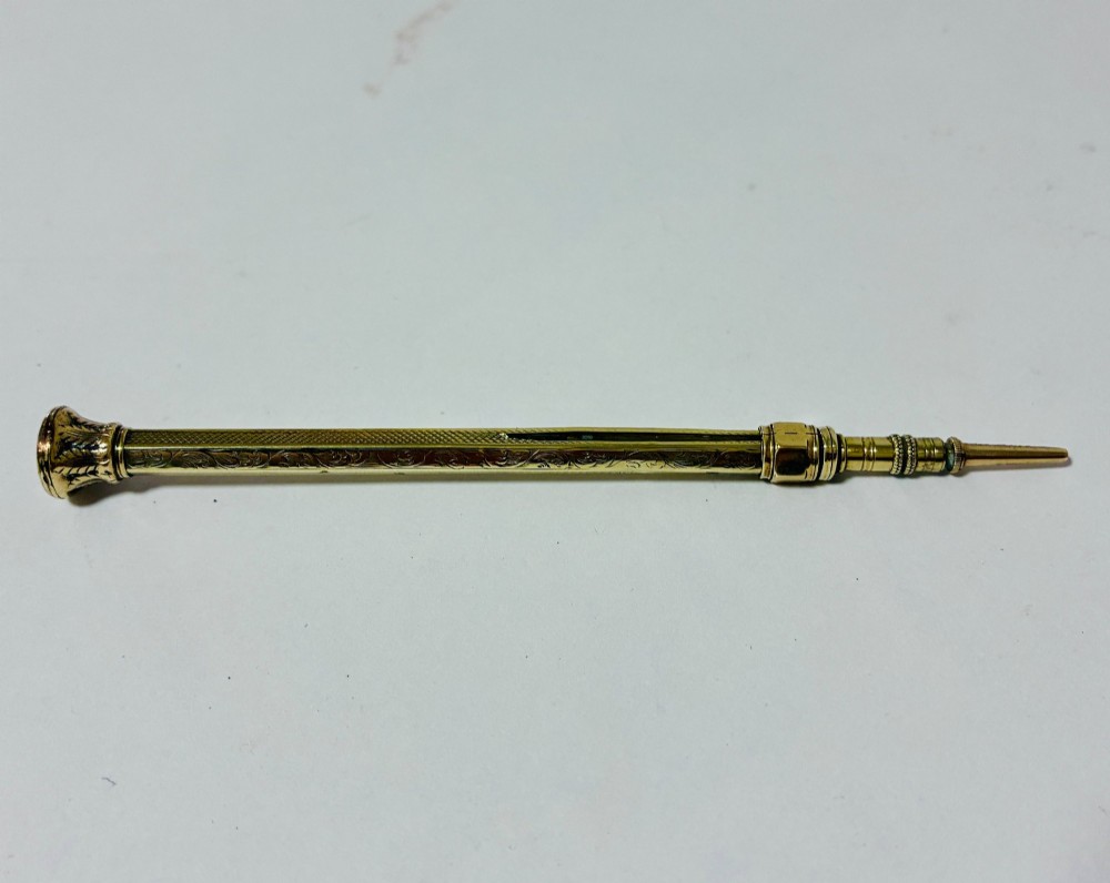 An antique gold propelling pencil featuring detailed scroll engraving along its slender hexagonal barrel and a polished tip.