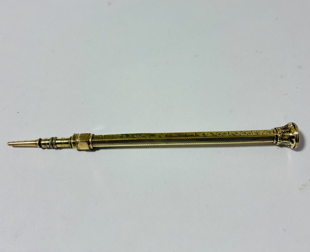 An antique gold propelling pencil featuring detailed scroll engraving along its slender hexagonal barrel and a polished tip. View VI