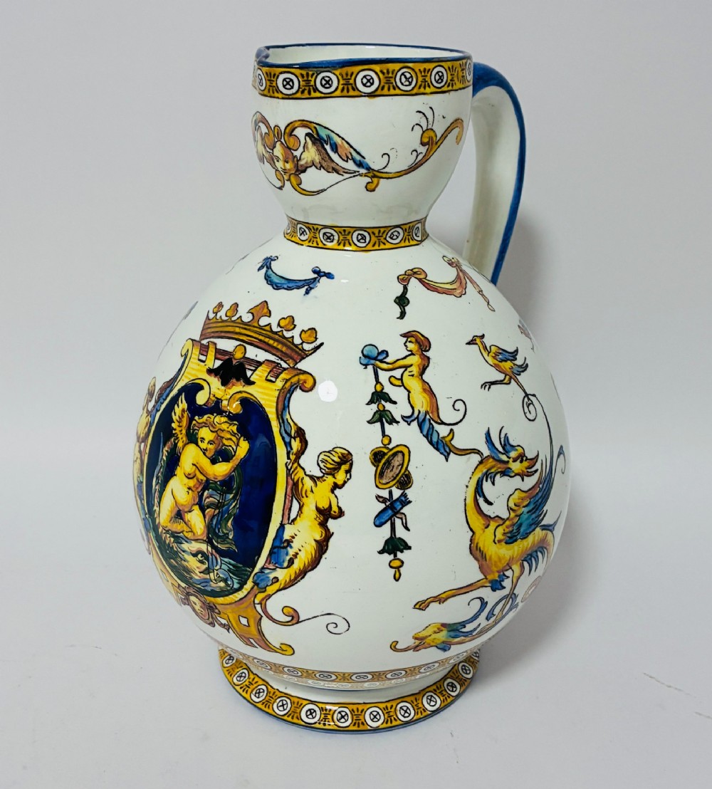 A white tin-glazed earthenware ewer decorated with hand-painted Renaissance grotesque figures, a central crest, and a blue handle.