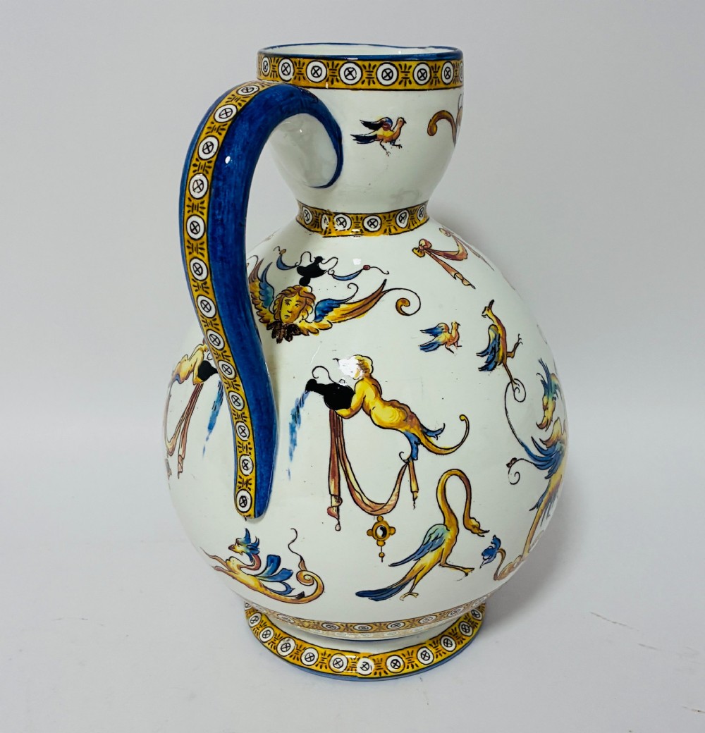 A white tin-glazed earthenware ewer decorated with hand-painted Renaissance grotesque figures, a central crest, and a blue handle. View IV
