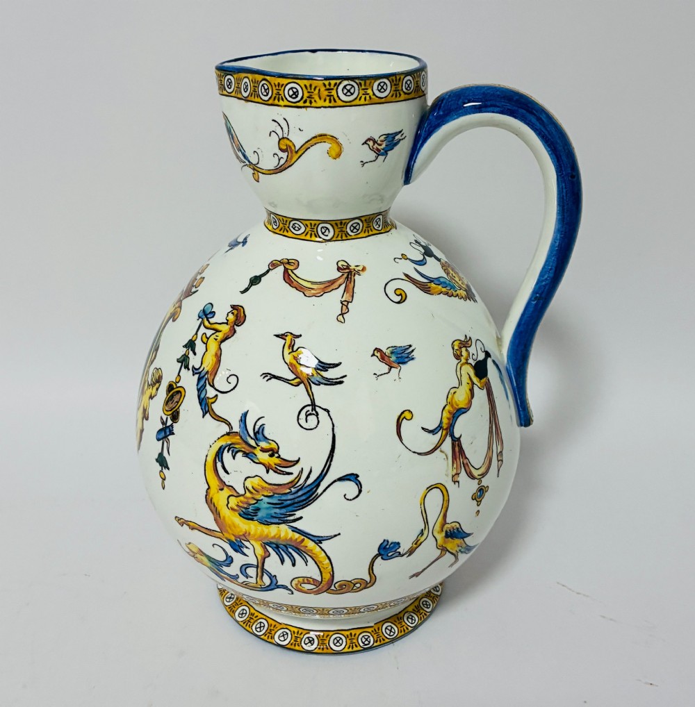A white tin-glazed earthenware ewer decorated with hand-painted Renaissance grotesque figures, a central crest, and a blue handle. View VI