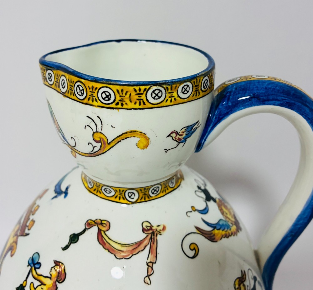 A white tin-glazed earthenware ewer decorated with hand-painted Renaissance grotesque figures, a central crest, and a blue handle. View VII