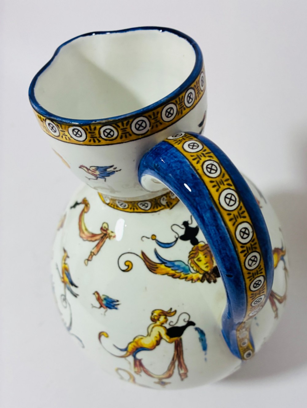 A white tin-glazed earthenware ewer decorated with hand-painted Renaissance grotesque figures, a central crest, and a blue handle. View IX