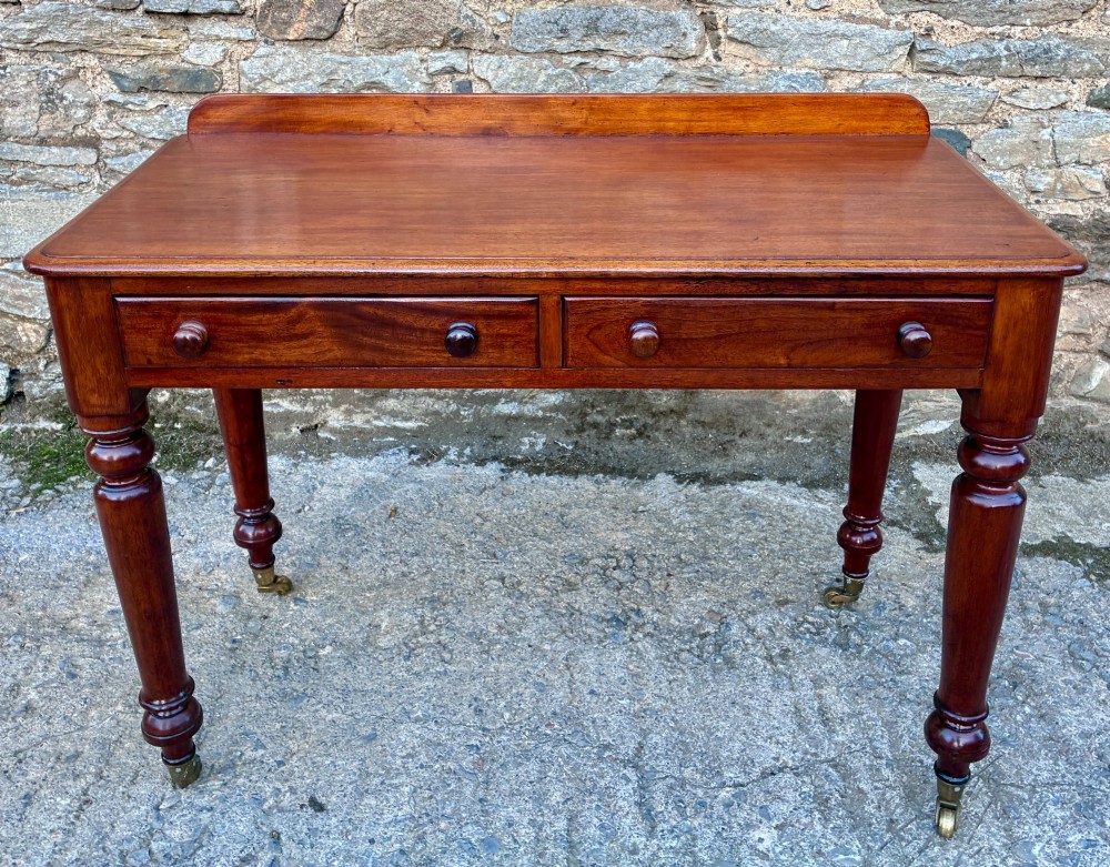 A Victorian mahogany side table featuring a low rear gallery, two drawers with wooden knob handles, turned legs, and brass castors.