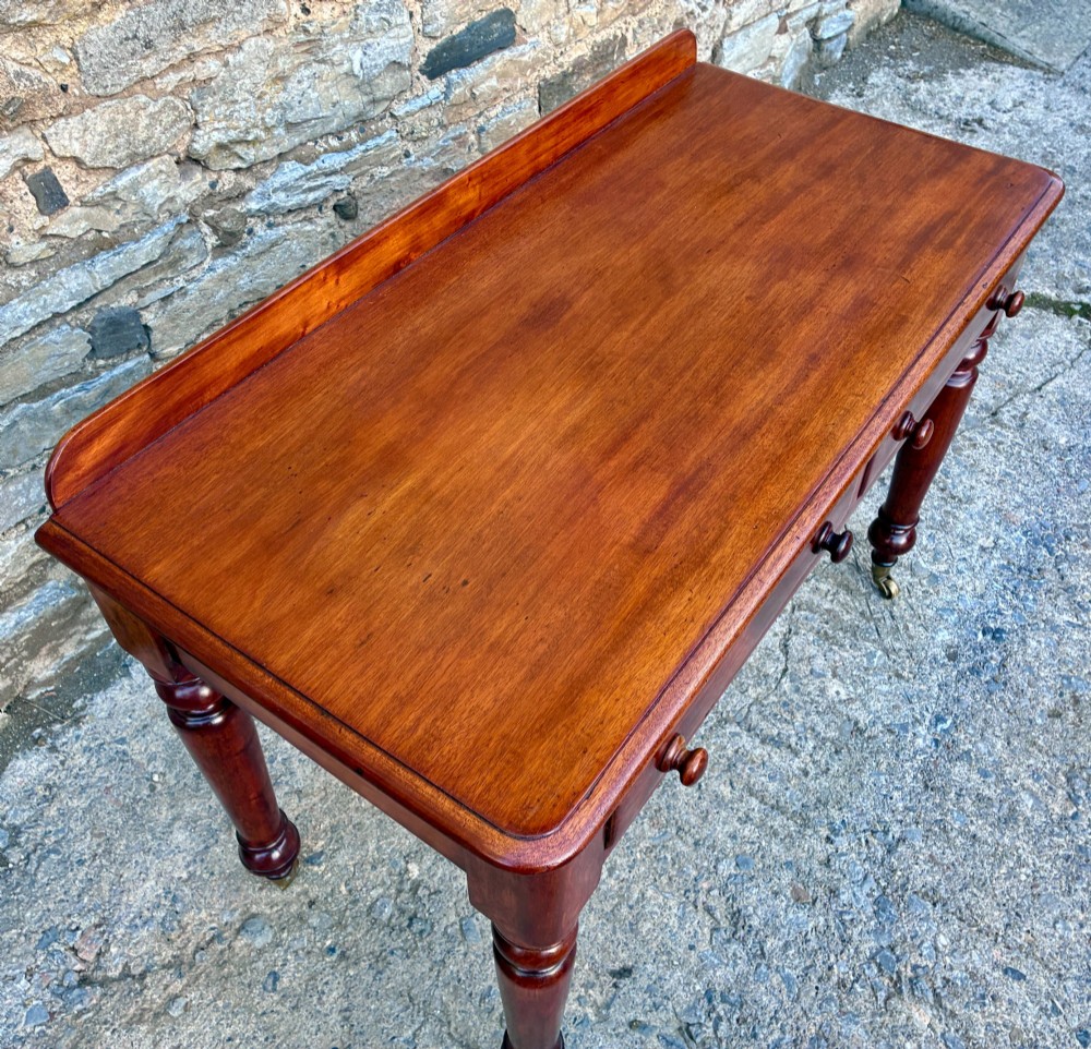 A Victorian mahogany side table featuring a low rear gallery, two drawers with wooden knob handles, turned legs, and brass castors. View IV