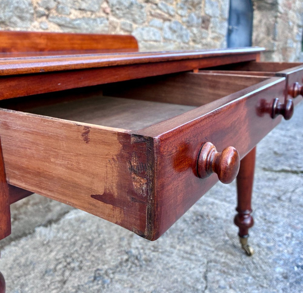 A Victorian mahogany side table featuring a low rear gallery, two drawers with wooden knob handles, turned legs, and brass castors. View VI