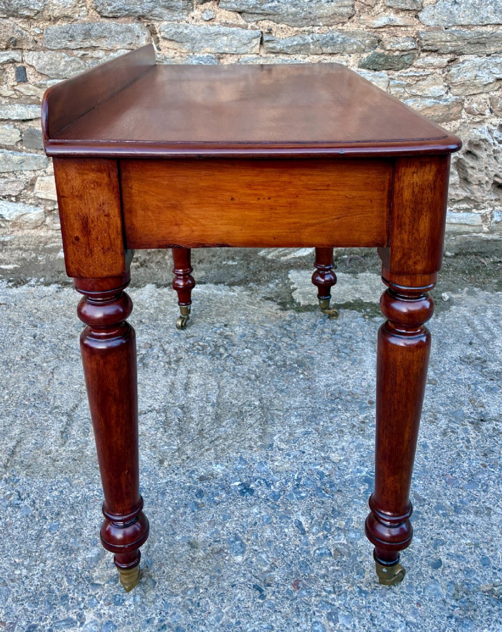 A Victorian mahogany side table featuring a low rear gallery, two drawers with wooden knob handles, turned legs, and brass castors. View X