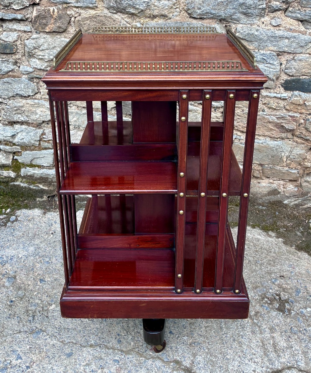 An Edwardian mahogany revolving bookcase featuring a square top with a brass gallery rail, slatted sides with brass studs, two tiers of shelving, and a central rotating mechanism on castors.