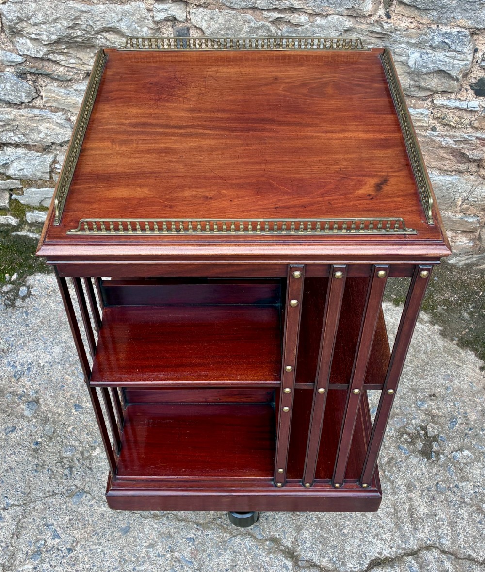 An Edwardian mahogany revolving bookcase featuring a square top with a brass gallery rail, slatted sides with brass studs, two tiers of shelving, and a central rotating mechanism on castors. View II