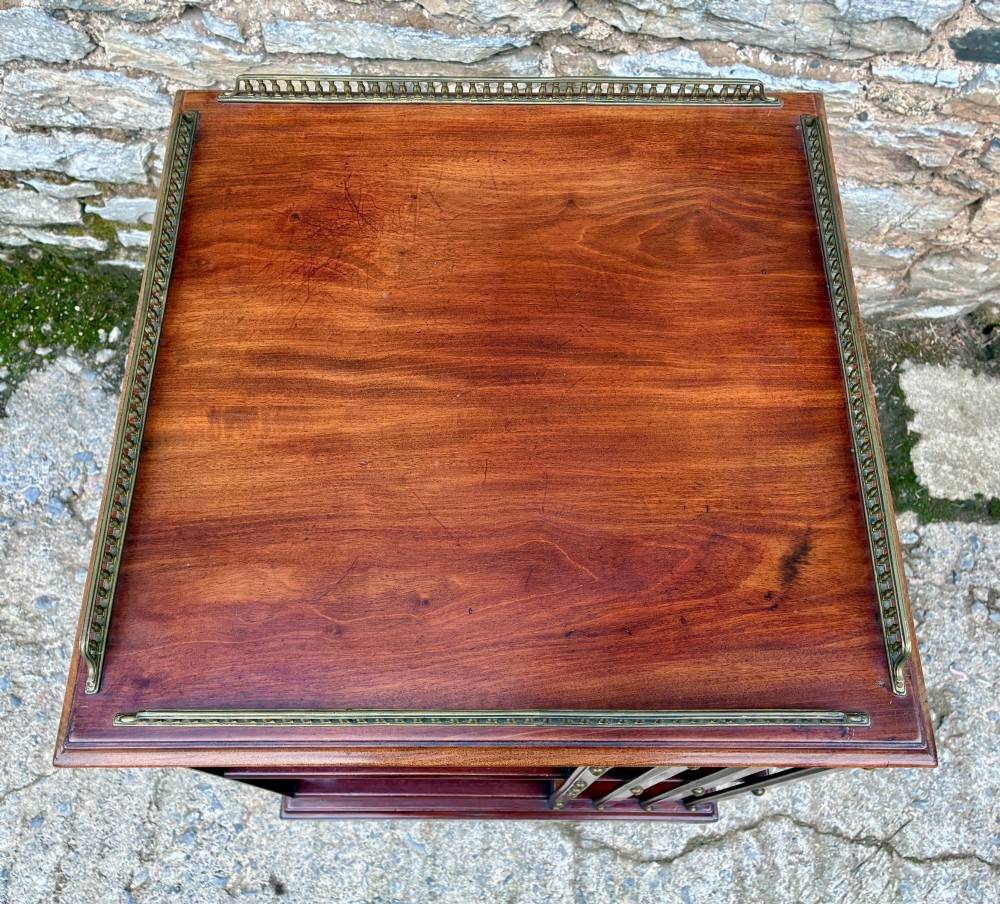 An Edwardian mahogany revolving bookcase featuring a square top with a brass gallery rail, slatted sides with brass studs, two tiers of shelving, and a central rotating mechanism on castors. View III