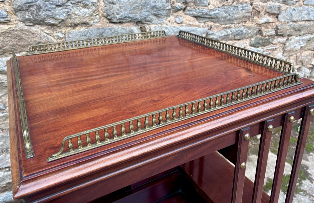 An Edwardian mahogany revolving bookcase featuring a square top with a brass gallery rail, slatted sides with brass studs, two tiers of shelving, and a central rotating mechanism on castors. View IV