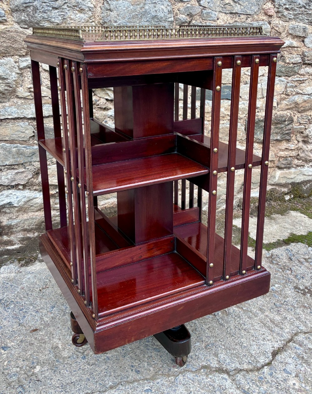 An Edwardian mahogany revolving bookcase featuring a square top with a brass gallery rail, slatted sides with brass studs, two tiers of shelving, and a central rotating mechanism on castors. View V