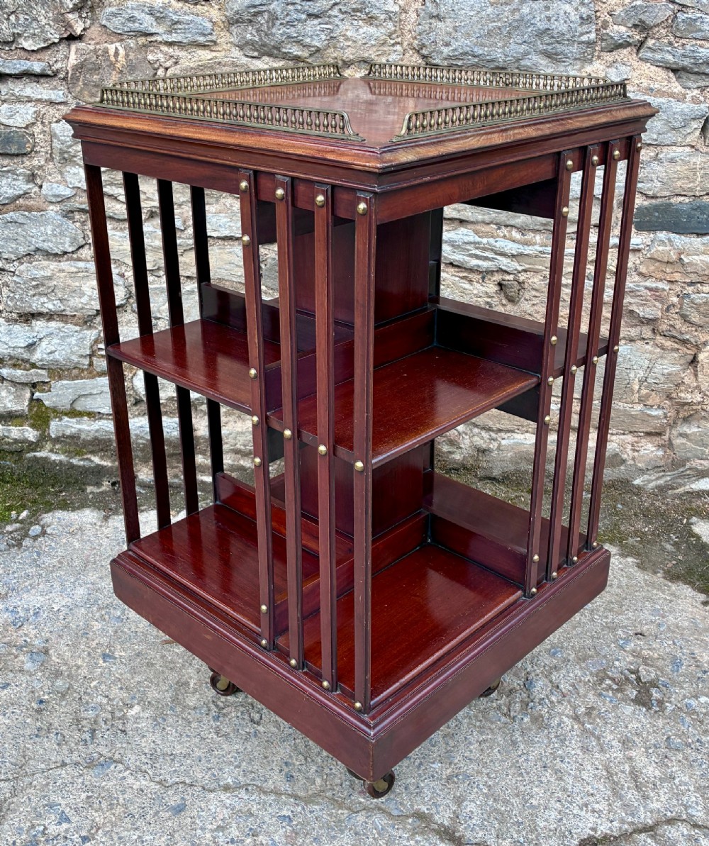 An Edwardian mahogany revolving bookcase featuring a square top with a brass gallery rail, slatted sides with brass studs, two tiers of shelving, and a central rotating mechanism on castors. View VI