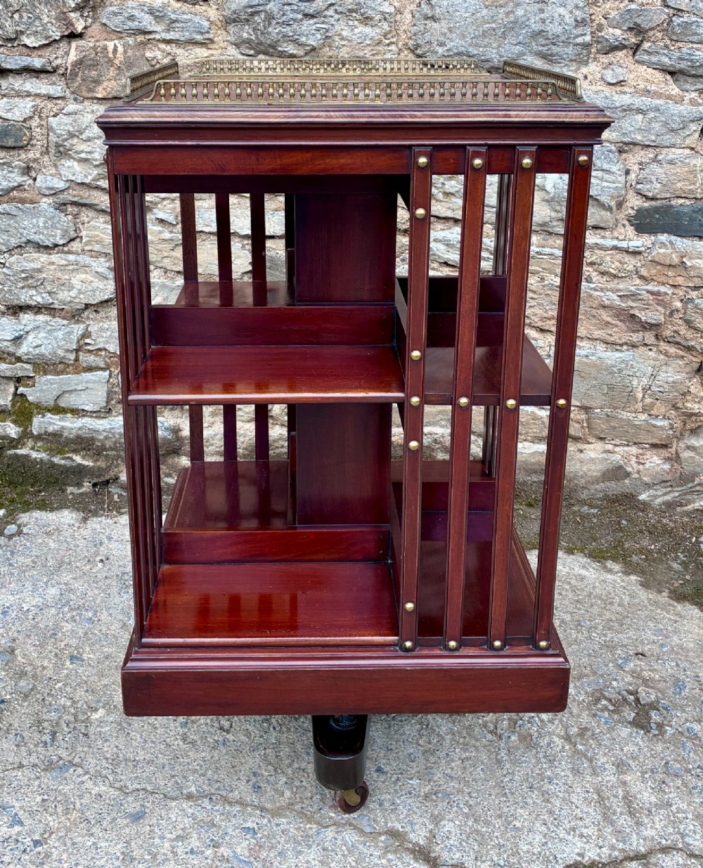 An Edwardian mahogany revolving bookcase featuring a square top with a brass gallery rail, slatted sides with brass studs, two tiers of shelving, and a central rotating mechanism on castors. View VII