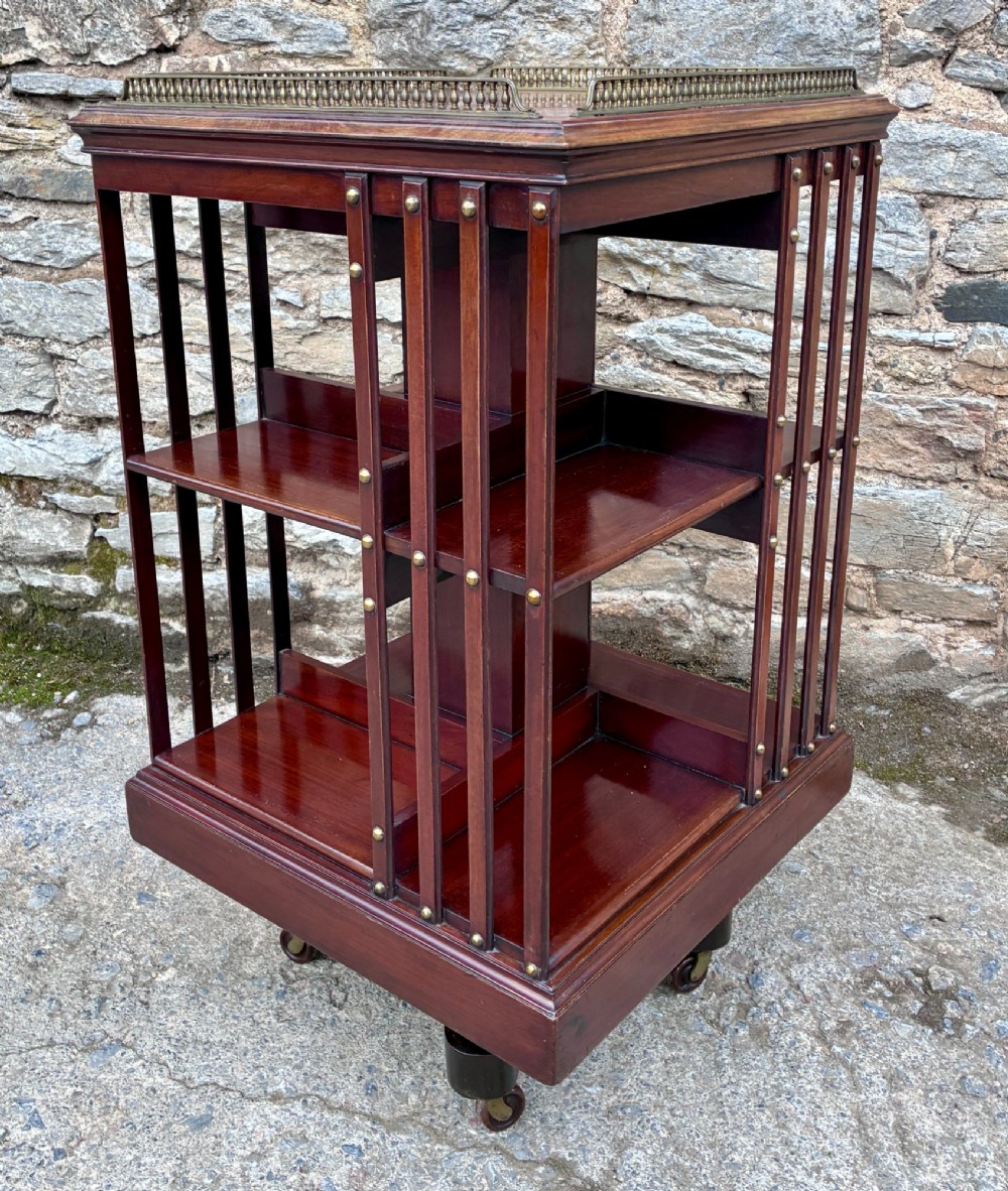 An Edwardian mahogany revolving bookcase featuring a square top with a brass gallery rail, slatted sides with brass studs, two tiers of shelving, and a central rotating mechanism on castors. View VIII