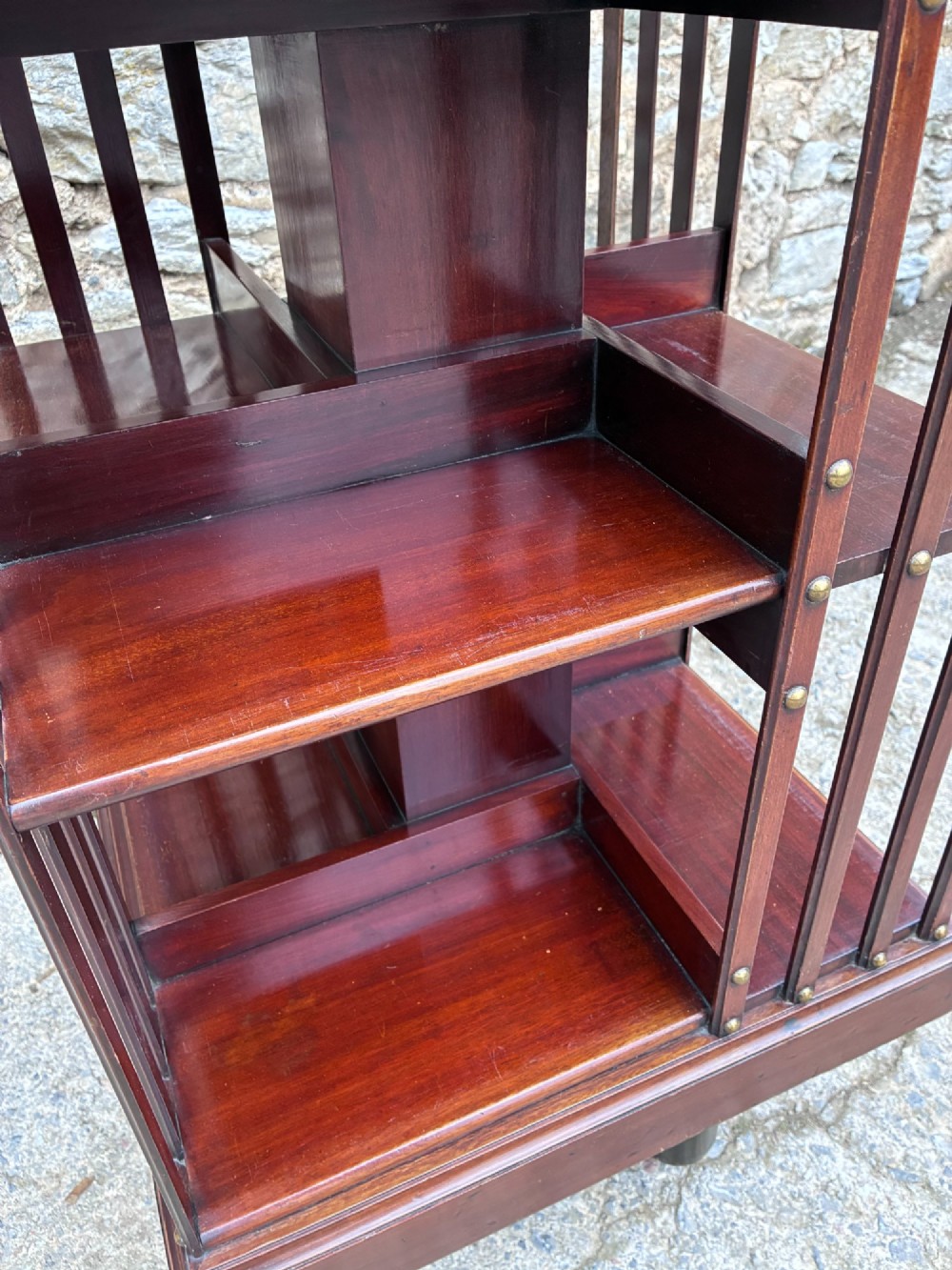 An Edwardian mahogany revolving bookcase featuring a square top with a brass gallery rail, slatted sides with brass studs, two tiers of shelving, and a central rotating mechanism on castors. View X