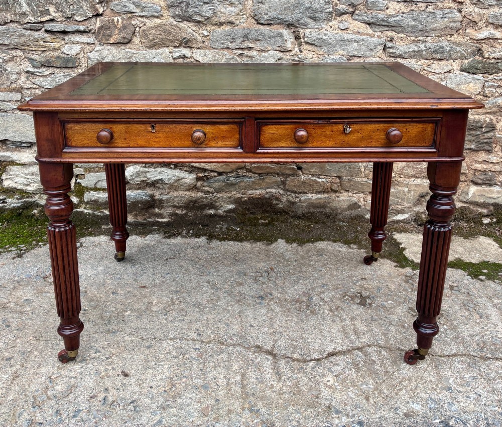 A Victorian mahogany writing table featuring a green leather top, two front drawers with turned wooden knobs, and fluted turned legs on brass castors. View II