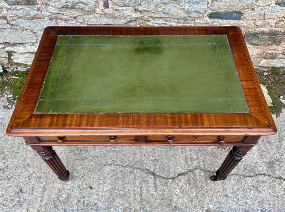 A Victorian mahogany writing table featuring a green leather top, two front drawers with turned wooden knobs, and fluted turned legs on brass castors. View III