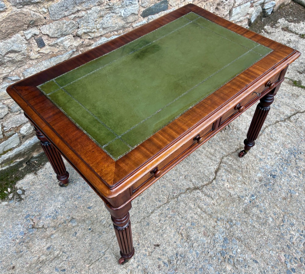 A Victorian mahogany writing table featuring a green leather top, two front drawers with turned wooden knobs, and fluted turned legs on brass castors. View IV