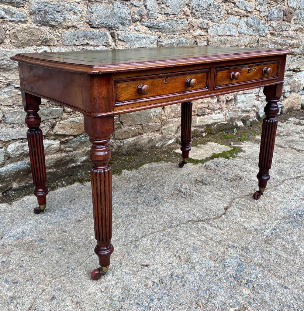 A Victorian mahogany writing table featuring a green leather top, two front drawers with turned wooden knobs, and fluted turned legs on brass castors. View V