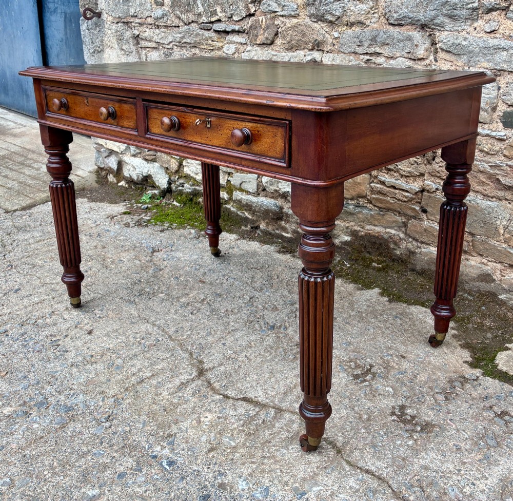 A Victorian mahogany writing table featuring a green leather top, two front drawers with turned wooden knobs, and fluted turned legs on brass castors. View VI