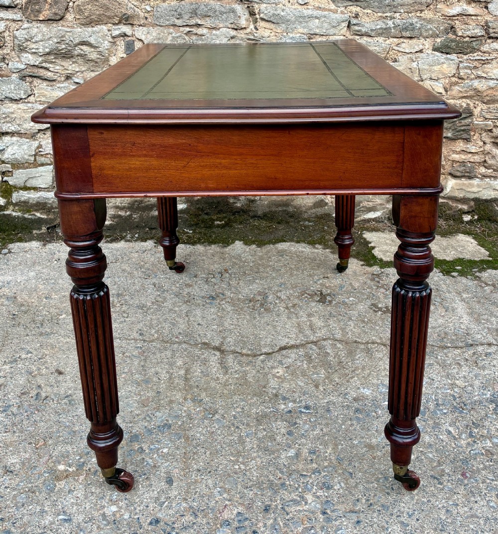 A Victorian mahogany writing table featuring a green leather top, two front drawers with turned wooden knobs, and fluted turned legs on brass castors. View XII