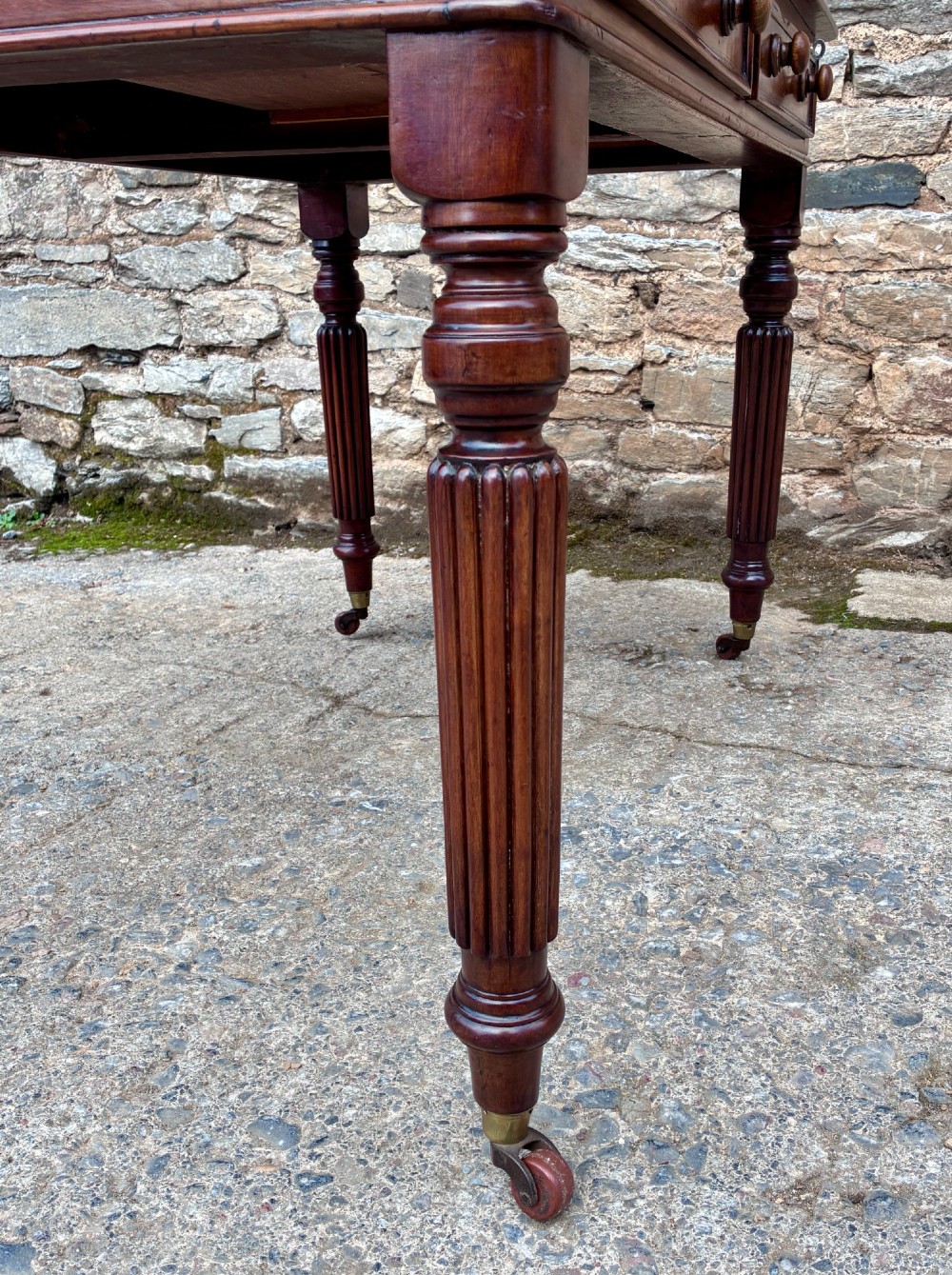 A Victorian mahogany writing table featuring a green leather top, two front drawers with turned wooden knobs, and fluted turned legs on brass castors. View XV