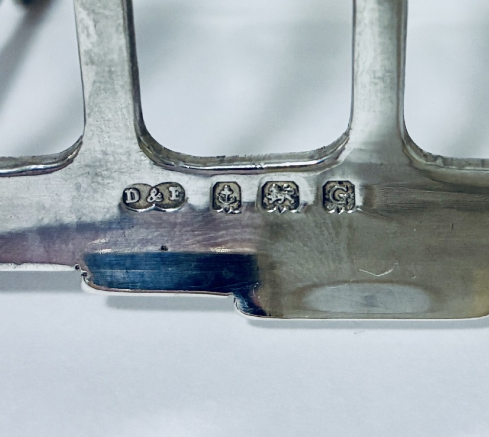 A Victorian sterling silver toast rack featuring four arched dividers, a central loop handle, and stamped hallmarks along the front base rail. View VII