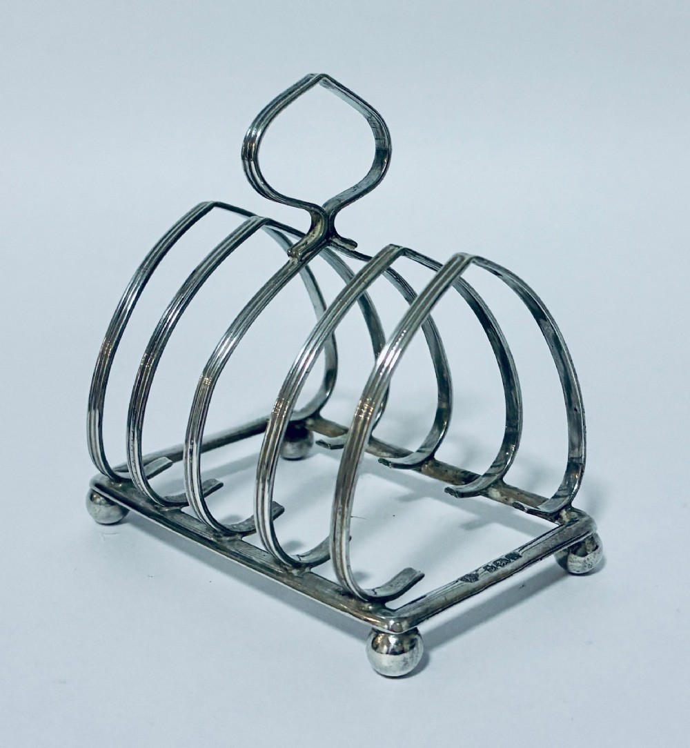 An antique silver toast rack featuring five arched divisions, a central loop handle, and a rectangular base supported by four ball feet.
