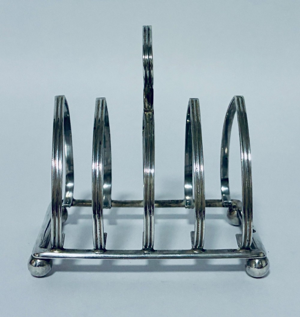 An antique silver toast rack featuring five arched divisions, a central loop handle, and a rectangular base supported by four ball feet. View II