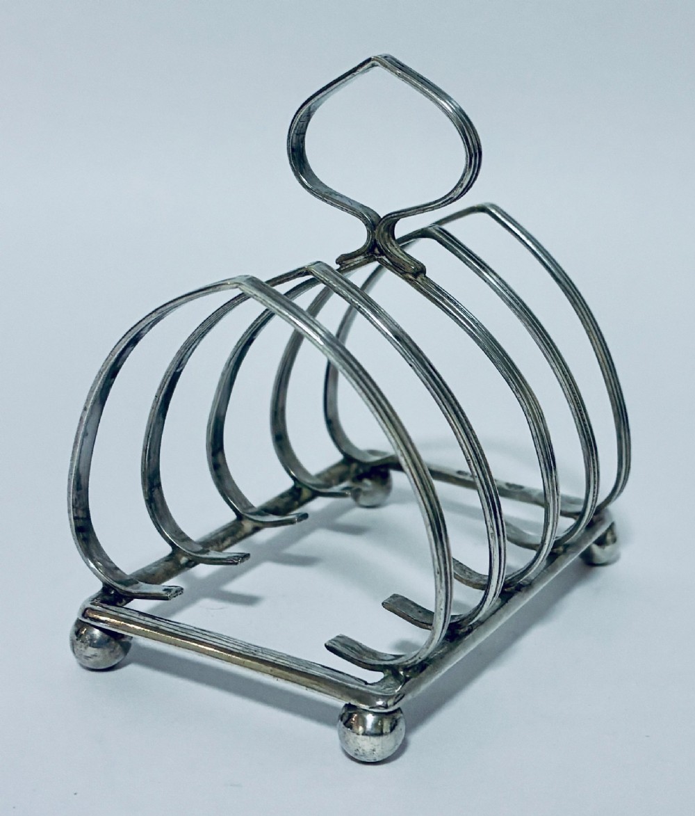 An antique silver toast rack featuring five arched divisions, a central loop handle, and a rectangular base supported by four ball feet. View III