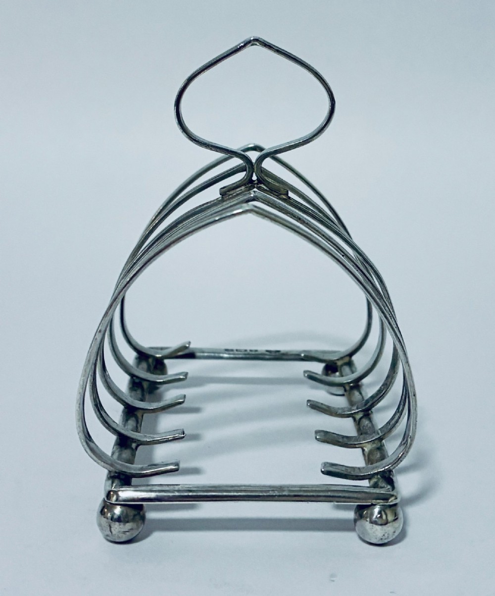 An antique silver toast rack featuring five arched divisions, a central loop handle, and a rectangular base supported by four ball feet. View IV
