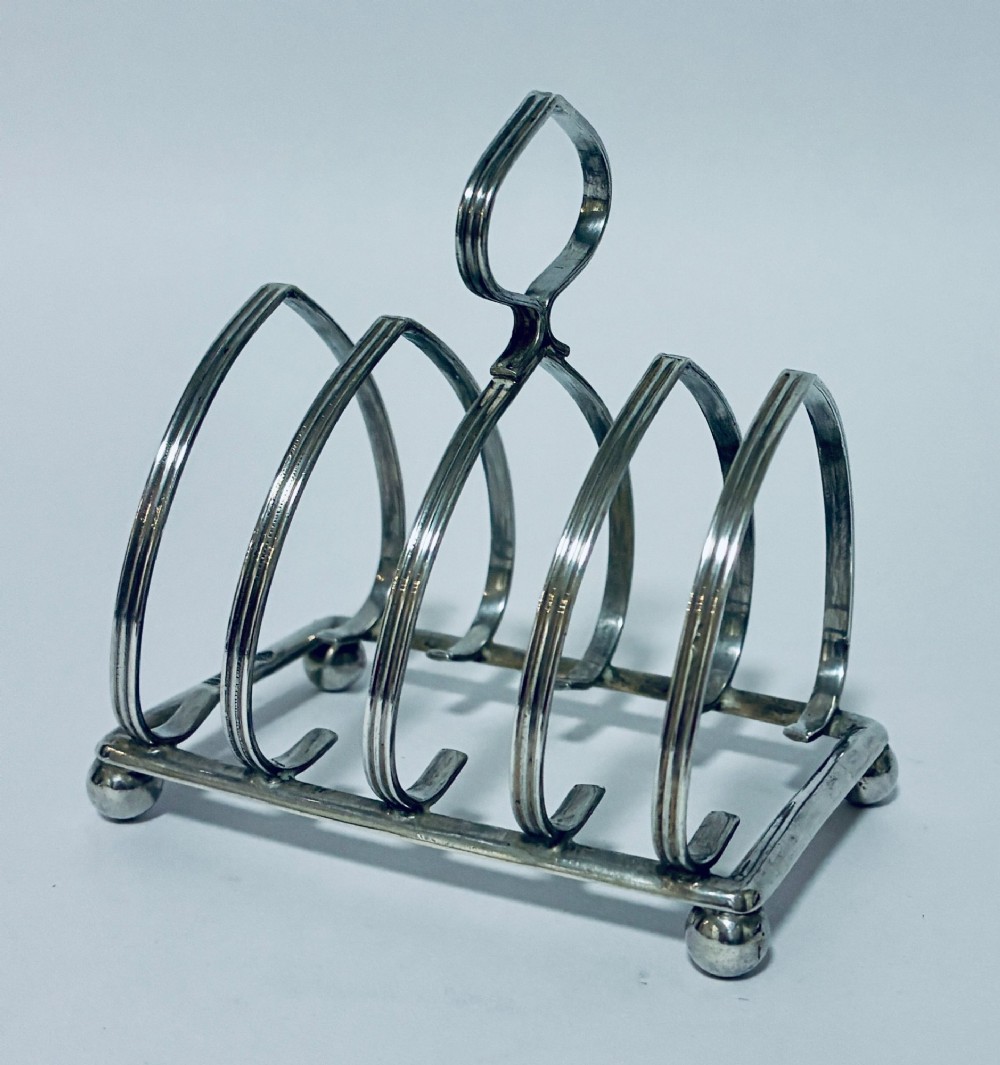 An antique silver toast rack featuring five arched divisions, a central loop handle, and a rectangular base supported by four ball feet. View V