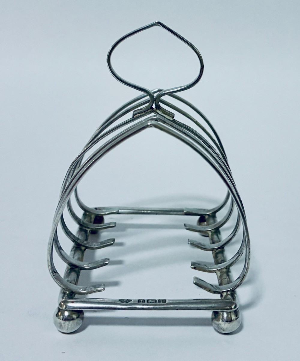 An antique silver toast rack featuring five arched divisions, a central loop handle, and a rectangular base supported by four ball feet. View VI