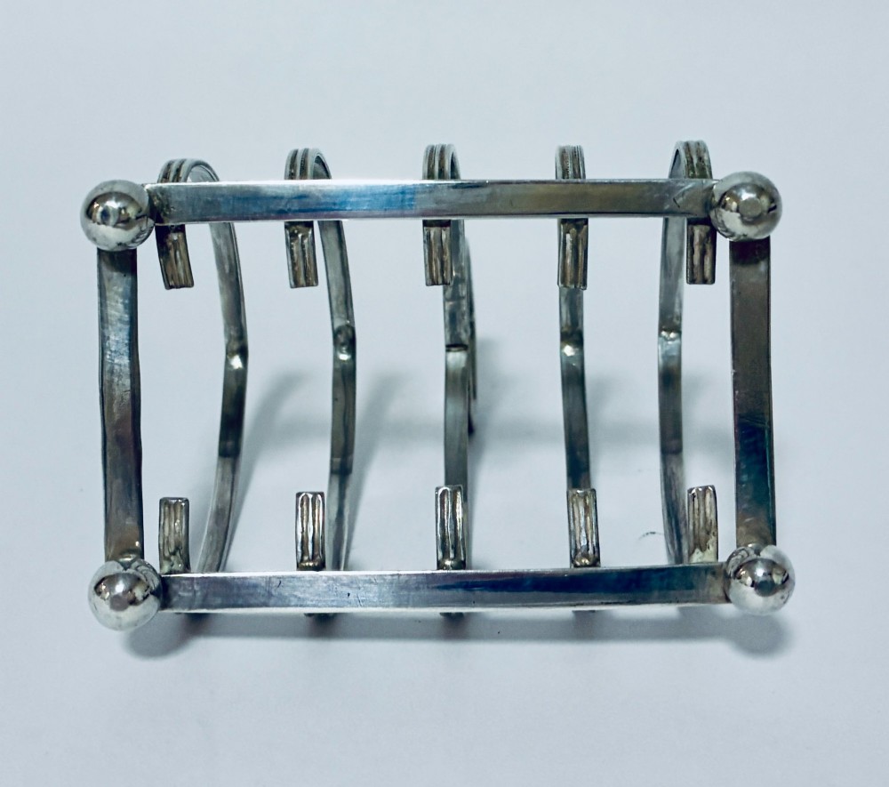 An antique silver toast rack featuring five arched divisions, a central loop handle, and a rectangular base supported by four ball feet. View IX