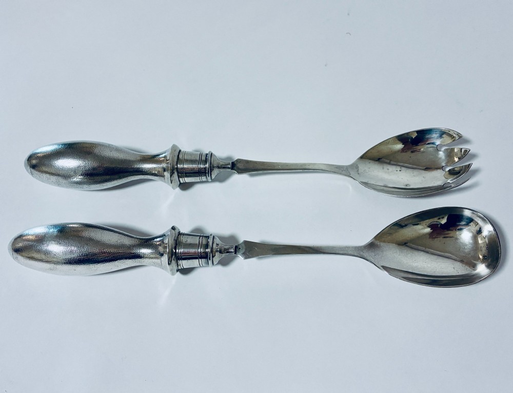 A pair of antique silver salad servers displaying planished hammered handles and polished metal serving ends.