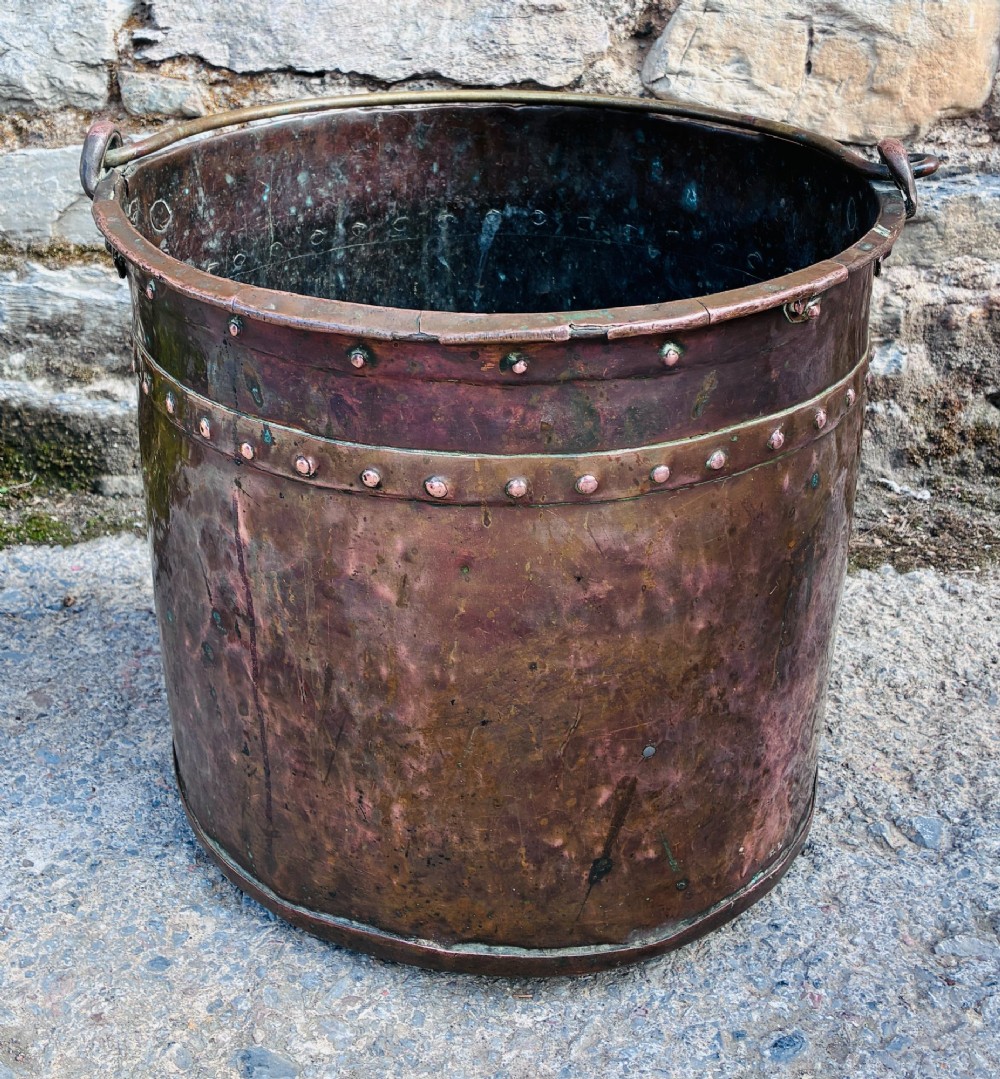 An antique cylindrical copper bucket featuring heavy copper rivets along the upper rim band and a metal swing handle.